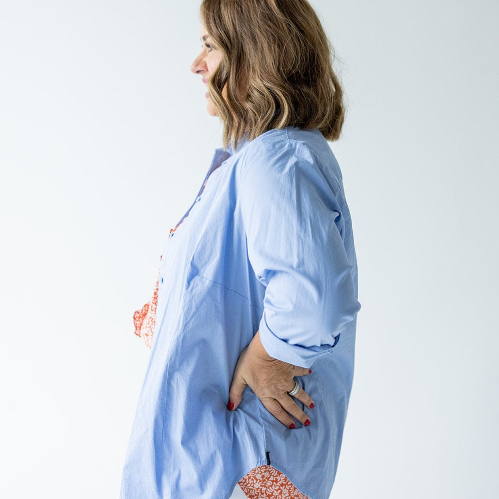 A woman in a CLASSIC BUTTON-UP BLOUSE IN AUTUMN SKY by Love Marlow made of lightweight Duratex fabric and white pants is standing in profile with her hand on her hip.