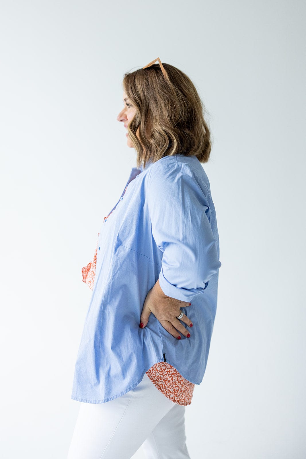 A woman in a CLASSIC BUTTON-UP BLOUSE IN AUTUMN SKY by Love Marlow made of lightweight Duratex fabric and white pants is standing in profile with her hand on her hip.