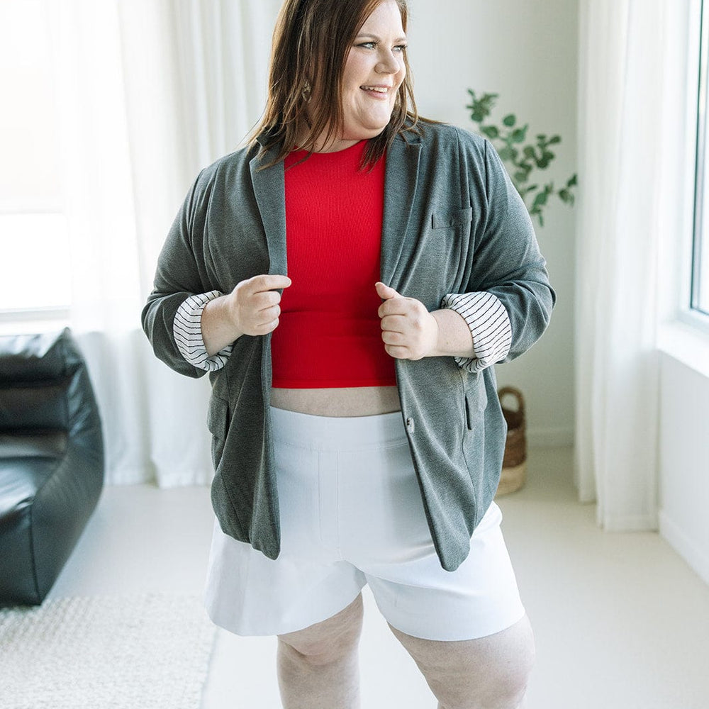 A person stands indoors wearing a red top, the CLASSIC BLAZER IN DARK GREY MELANGE from Love Marlow, white shorts, and black sandals, embodying a chic yet professional style. Sunglasses rest on top of their head as they look to the side.