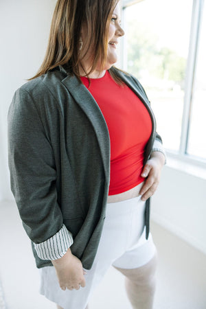 A person wearing Love Marlow's CLASSIC BLAZER IN DARK GREY MELANGE, paired with a red top and white shorts, stands near a window, smiling and touching their midsection, exuding professional elegance.