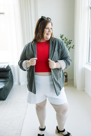 A person stands indoors wearing a red top, the CLASSIC BLAZER IN DARK GREY MELANGE from Love Marlow, white shorts, and black sandals, embodying a chic yet professional style. Sunglasses rest on top of their head as they look to the side.