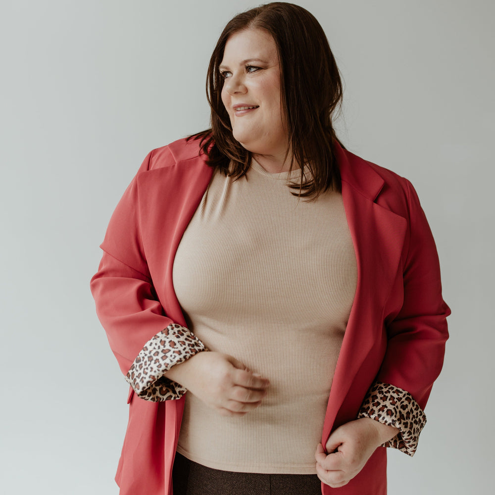 A person with long hair wearing an Oddi CLASSIC BLAZER IN LIPSTICK and a beige top stands against a gray background, perfectly complementing their lipstick color.