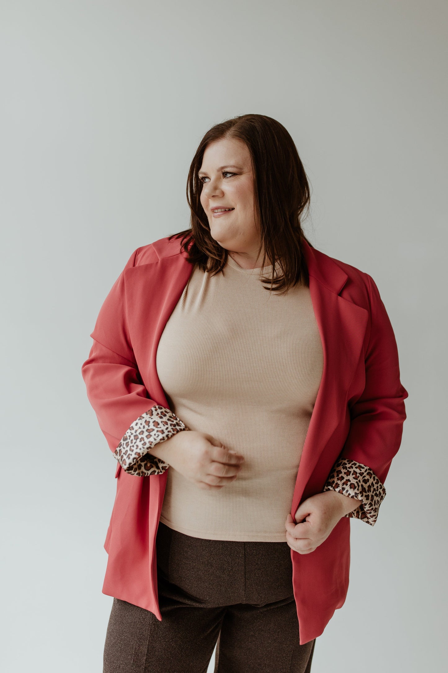 A person with long hair wearing an Oddi CLASSIC BLAZER IN LIPSTICK and a beige top stands against a gray background, perfectly complementing their lipstick color.