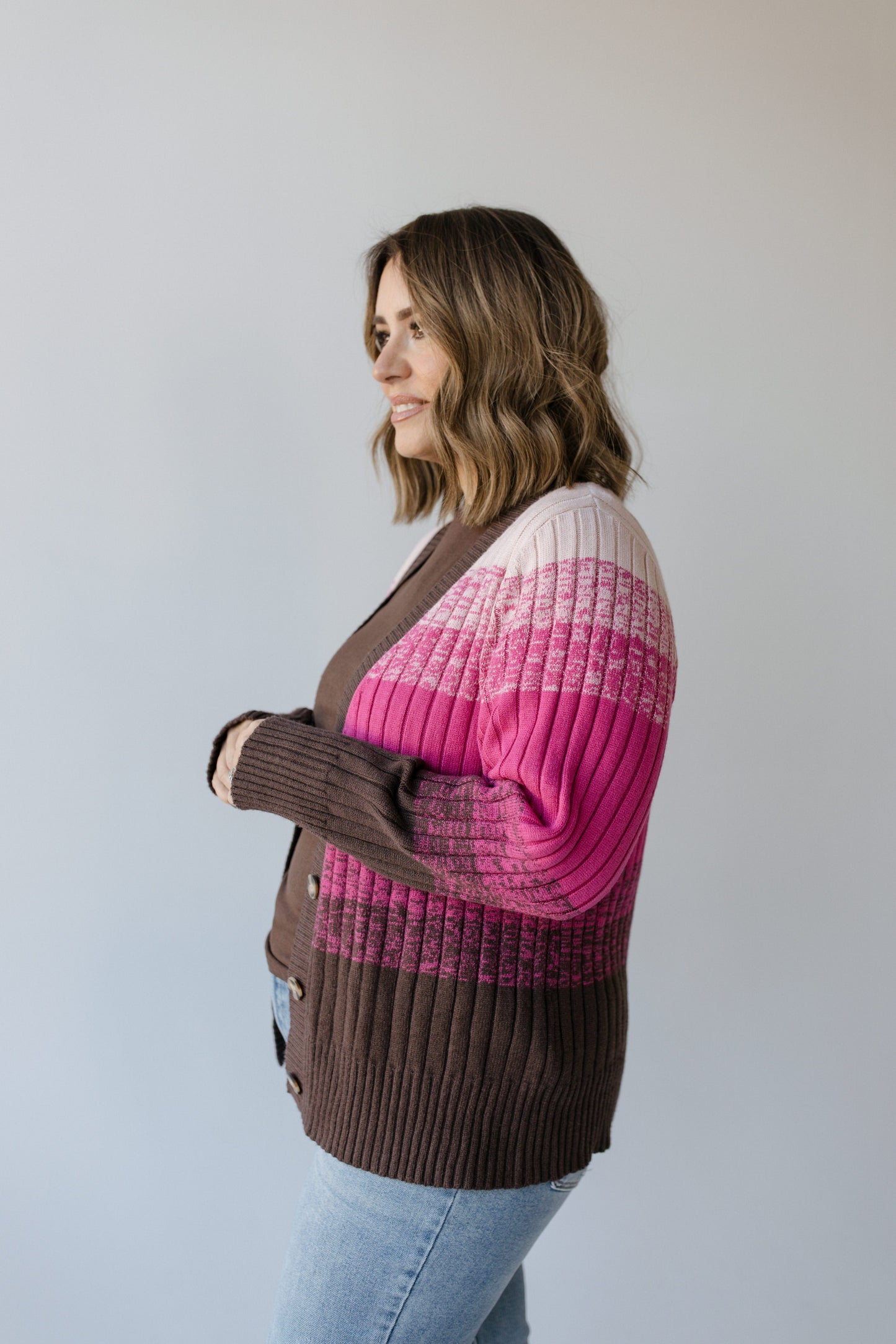 A person wearing the Yesta CLASSIC CARDIGAN WITH COLOR FADE IN PINK, showcasing pink, brown, and white stripes, stands sideways against a plain background.