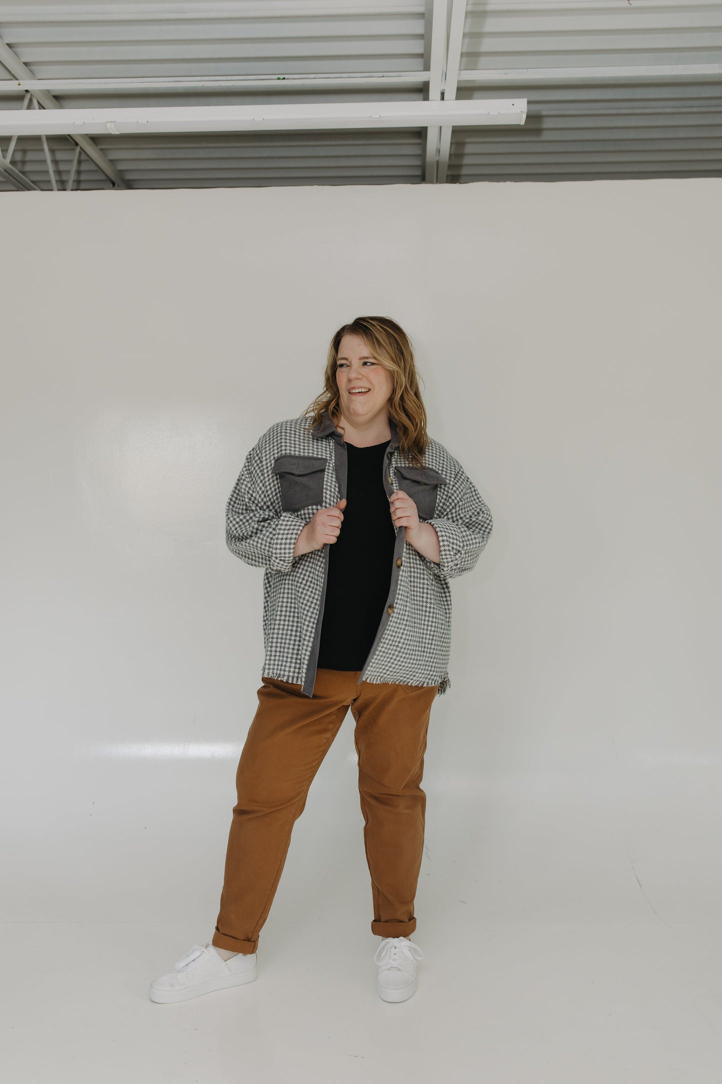 Person wearing a checkered jacket and brown pants standing against a white wall.