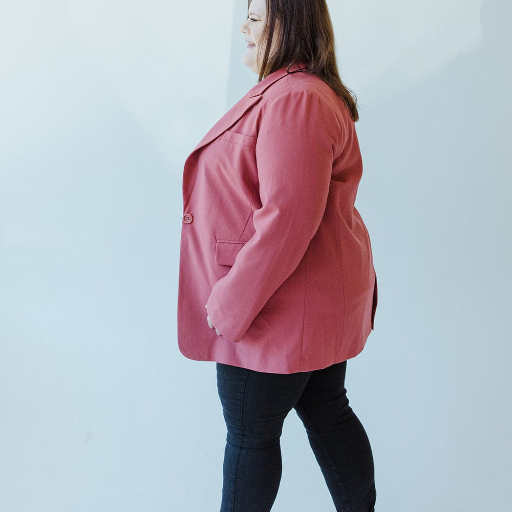 A person with long hair stands sideways, wearing a pink Yesta CLASSIC NOTCHED BLAZER paired with black pants and shoes, creating a sophisticated silhouette against the plain light background.