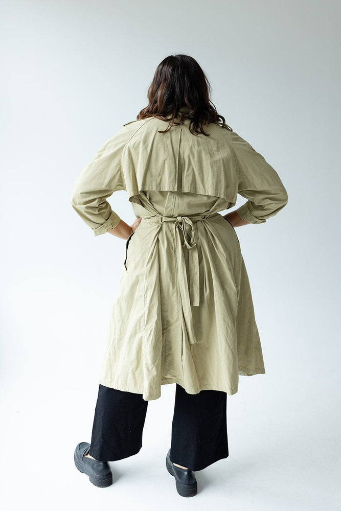 A person with long hair, wearing the Yesta CLASSIC TRENCH COAT crafted from water-resistant fabric with double-breasted buttons, stands facing away in a minimalistic setting.