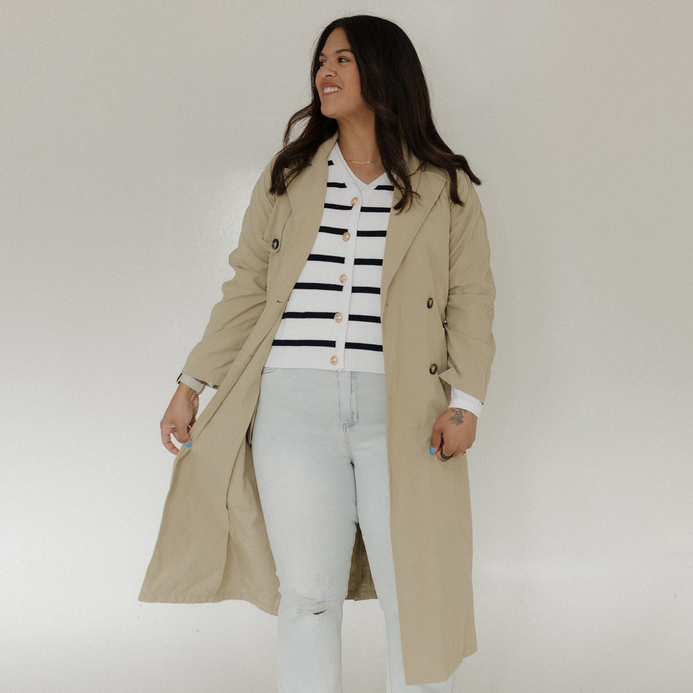 Woman wearing a beige trench coat over a striped shirt with white pants on a plain background