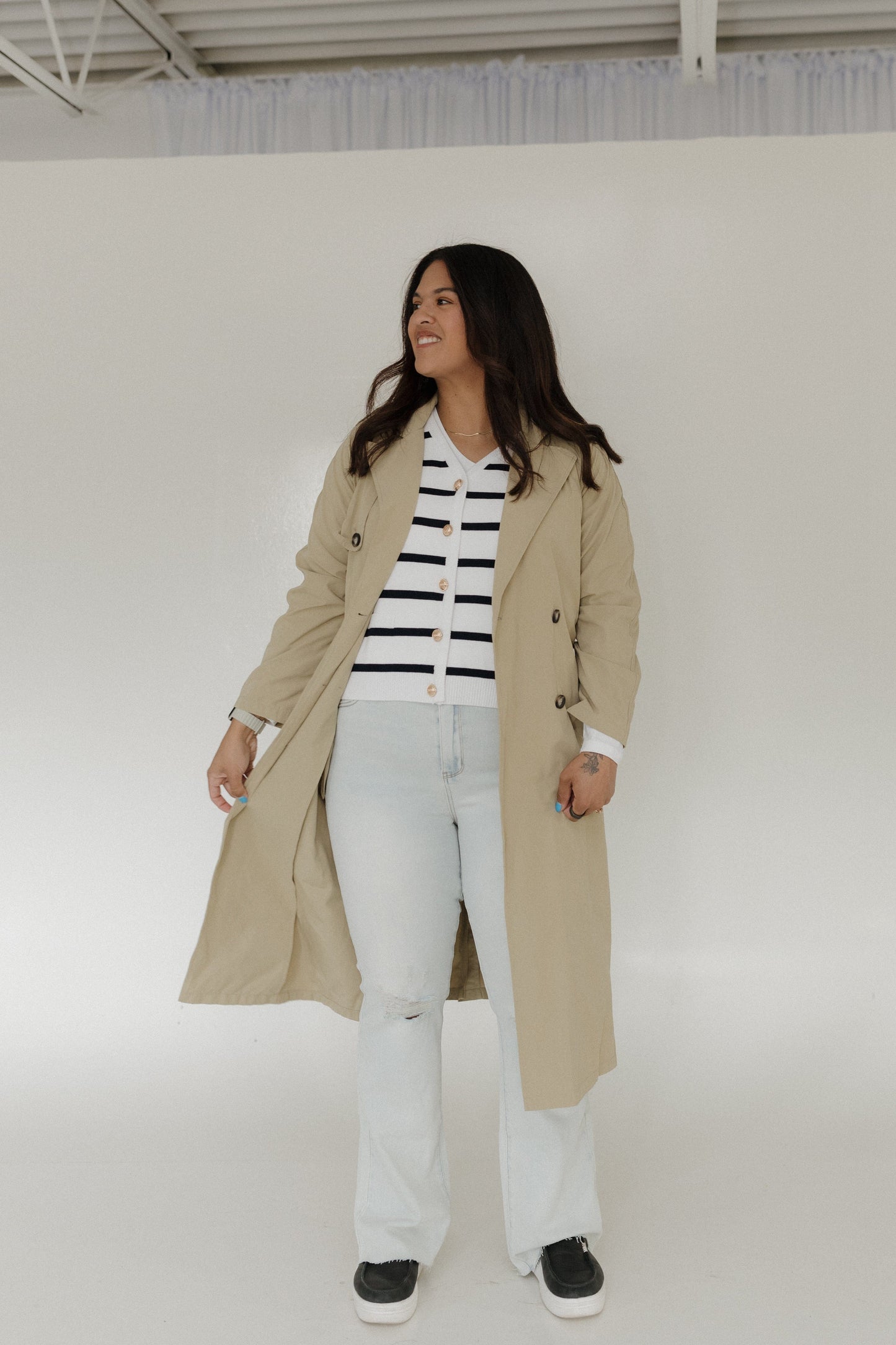 Woman wearing a beige trench coat over a striped shirt with white pants on a plain background