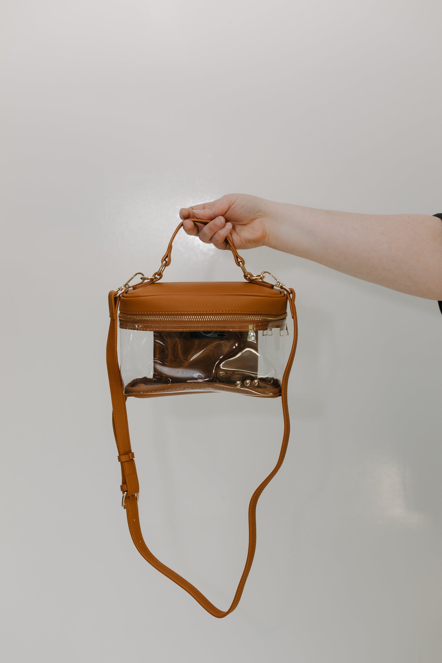 Clear handbag with brown leather handle and strap held by a person against a plain background