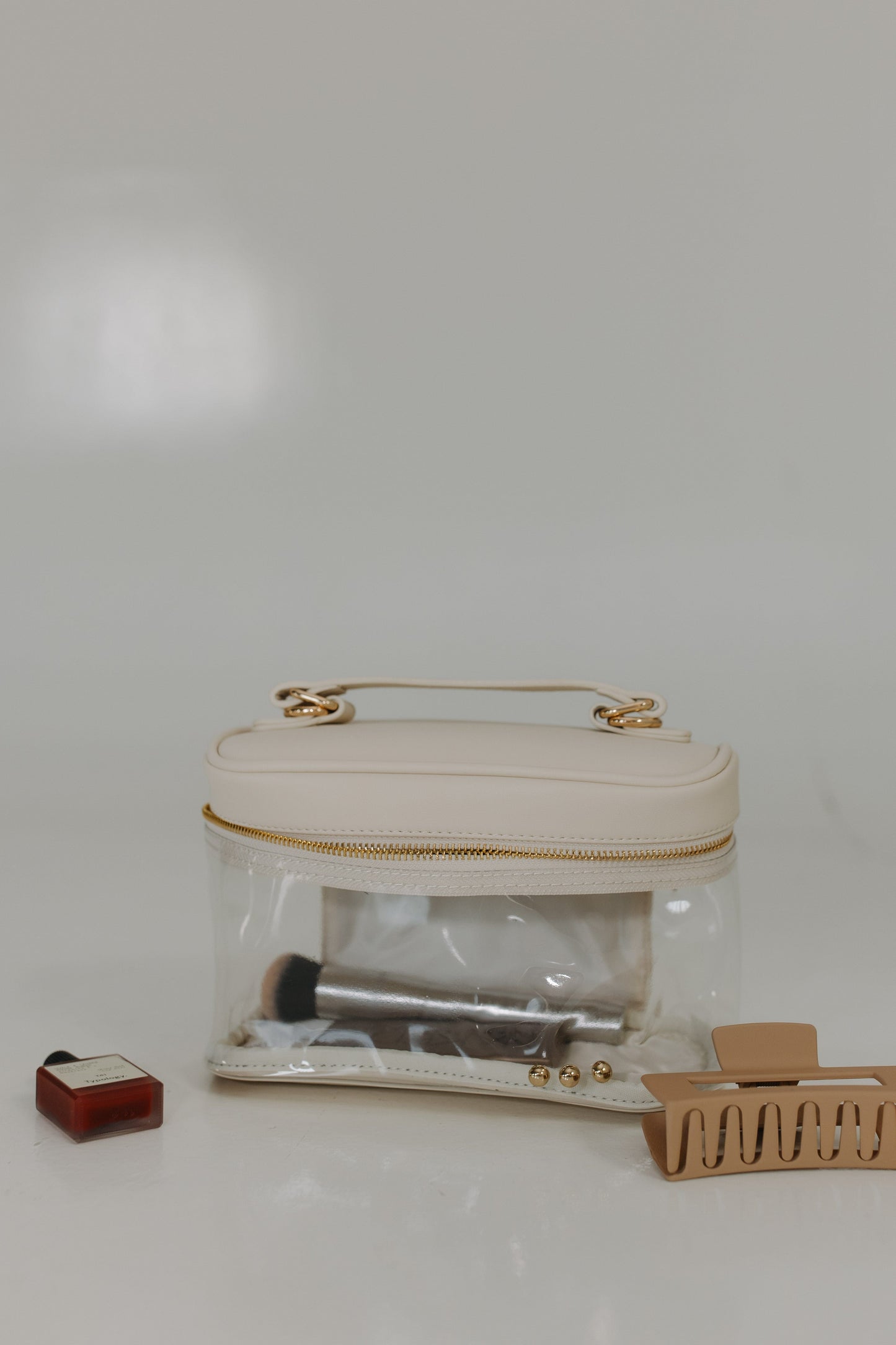 Clear plastic bag with gold handle, makeup items, and a hair clip on a white background