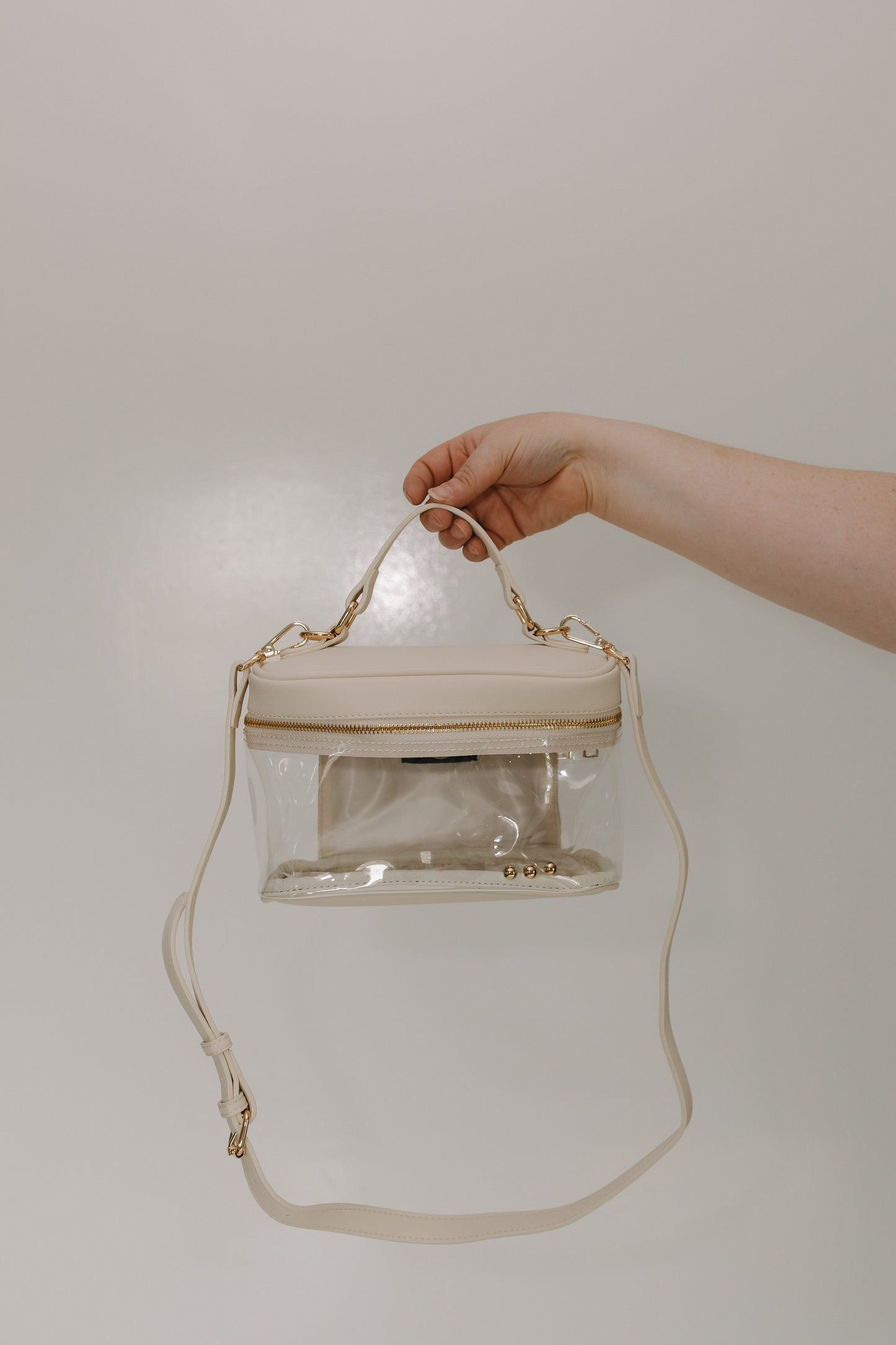 Clear handbag with gold accents held by a hand against a plain background