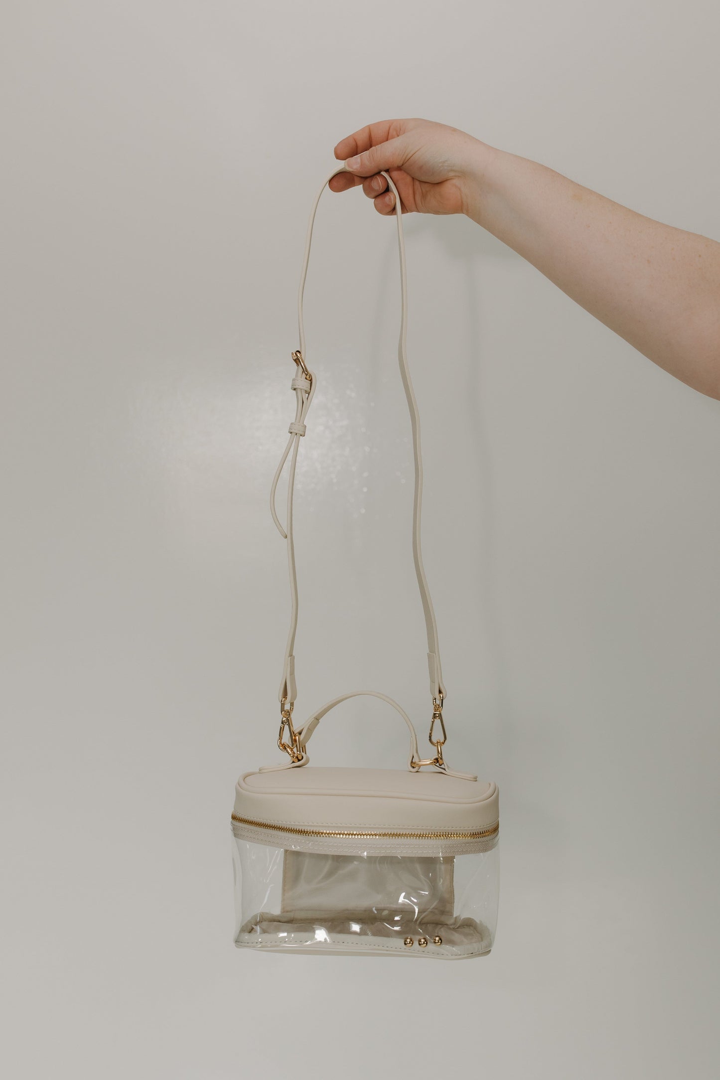 Clear handbag with gold accents held by a hand against a plain background