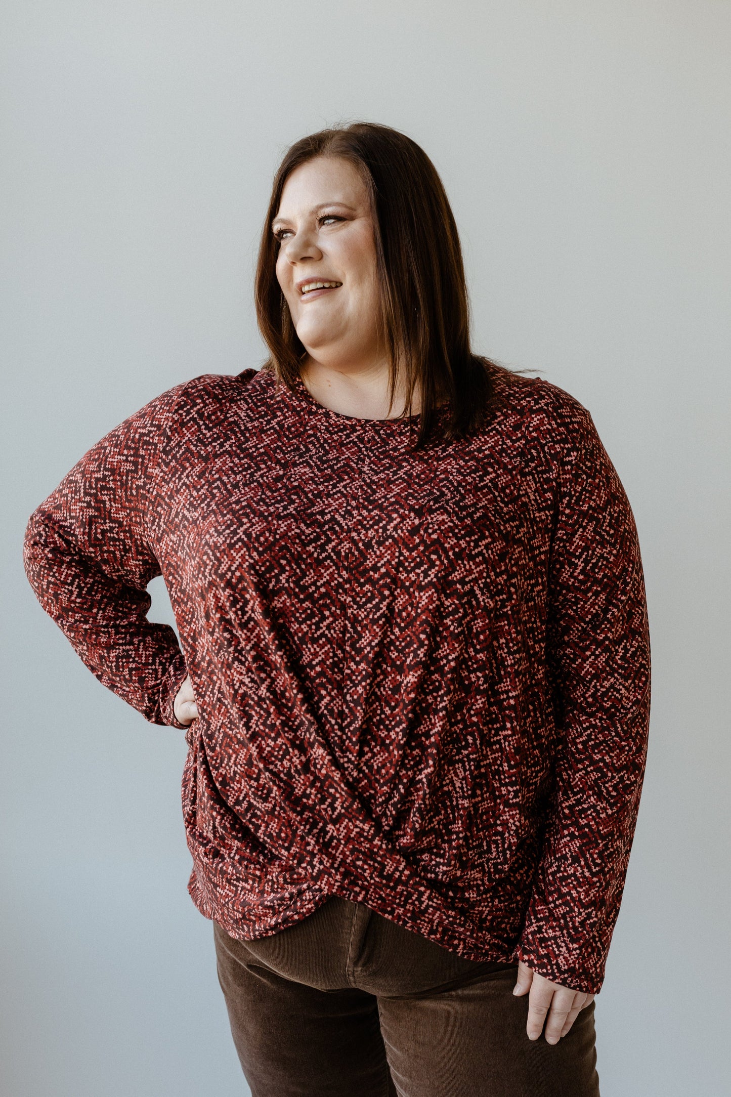 A person wearing a colorful pixelated red sweater with long sleeves and brown pants stands against a plain background, smiling while looking to the side.