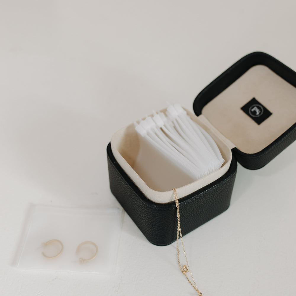 Black jewelry box with white interior, containing a necklace and two rings on a light gray background.
