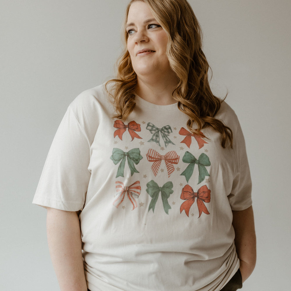 Person wearing a COQUETTE CHRISTMAS BOW GRAPHIC TEE with red and green festive bow designs, looking to the side, hands in their pockets against a plain background.