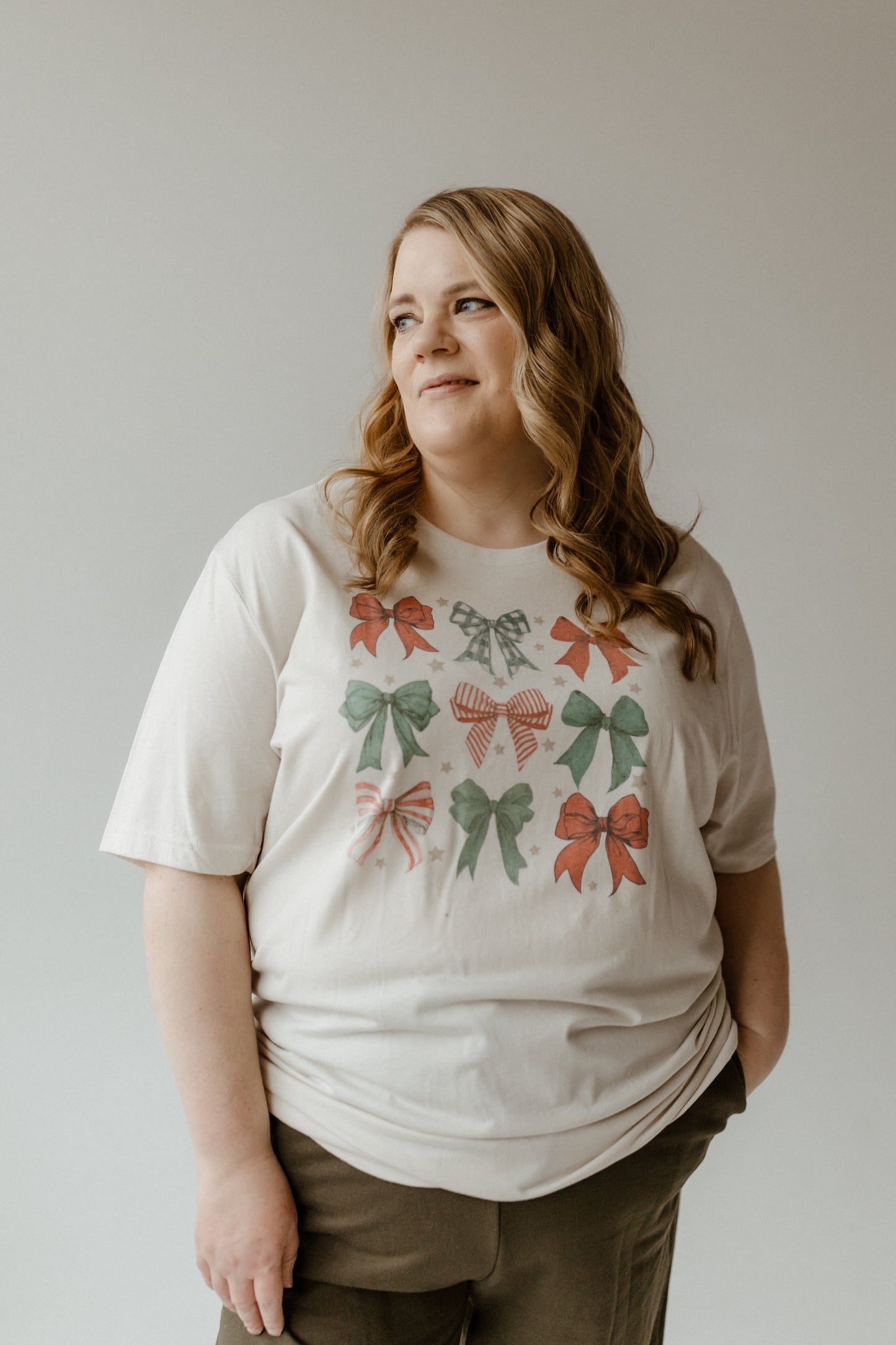 Person wearing a COQUETTE CHRISTMAS BOW GRAPHIC TEE with red and green festive bow designs, looking to the side, hands in their pockets against a plain background.