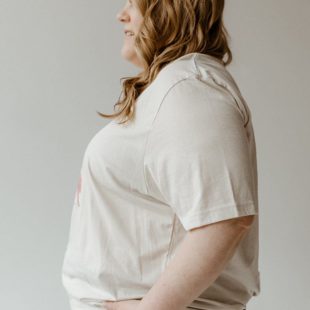 A person with long hair, wearing a festive bow graphic tee and dark pants, stands in profile with hands in pockets against a neutral background.