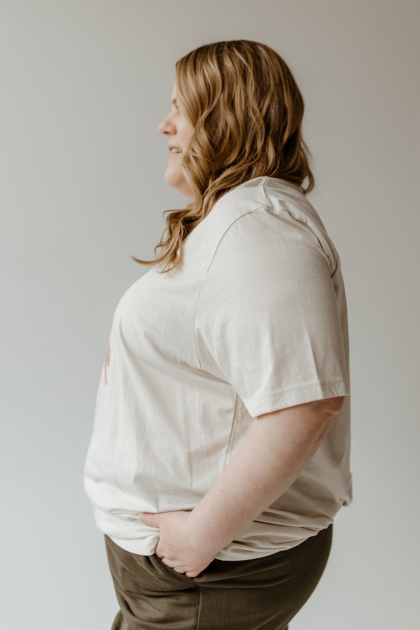 A person with long hair, wearing a festive bow graphic tee and dark pants, stands in profile with hands in pockets against a neutral background.