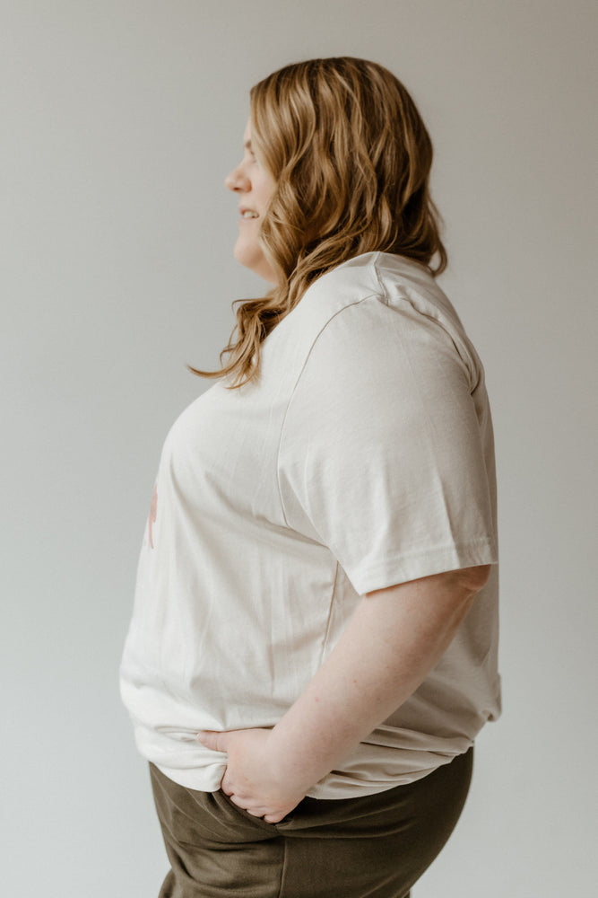 A person with long hair stands with their back to the camera, wearing a COQUETTE CHRISTMAS BOW GRAPHIC TEE in super soft fabric and dark pants against a plain background.