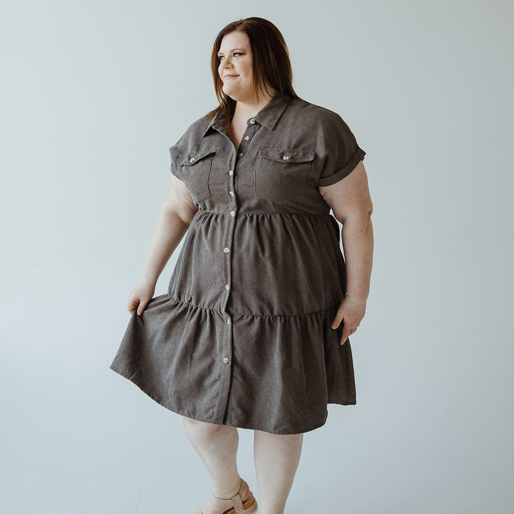 A person stands on a light background, wearing a short-sleeve, knee-length gray button-front dress paired with sandals.