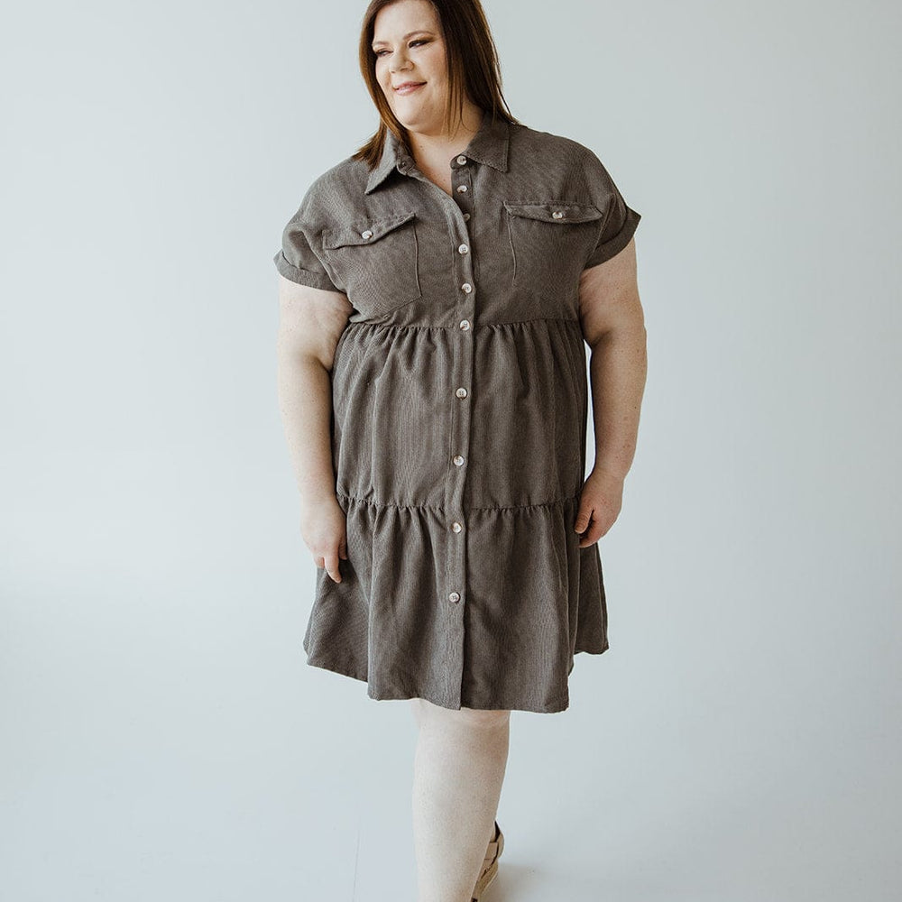 A person wears a gray, knee-length button-up dress featuring a tiered skirt, standing gracefully against a plain backdrop.