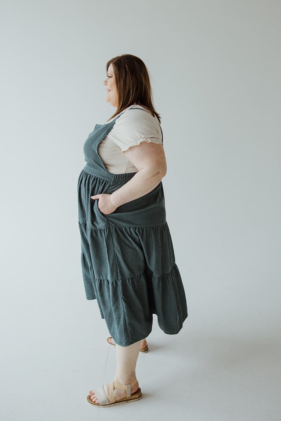 A person standing in profile view, clad in a white t-shirt under a Love Marlow CORDUROY TIERED JUMPER IN BLUE SPRUCE. The individual has one hand in a pocket and is looking forward. The background is plain and light-colored.