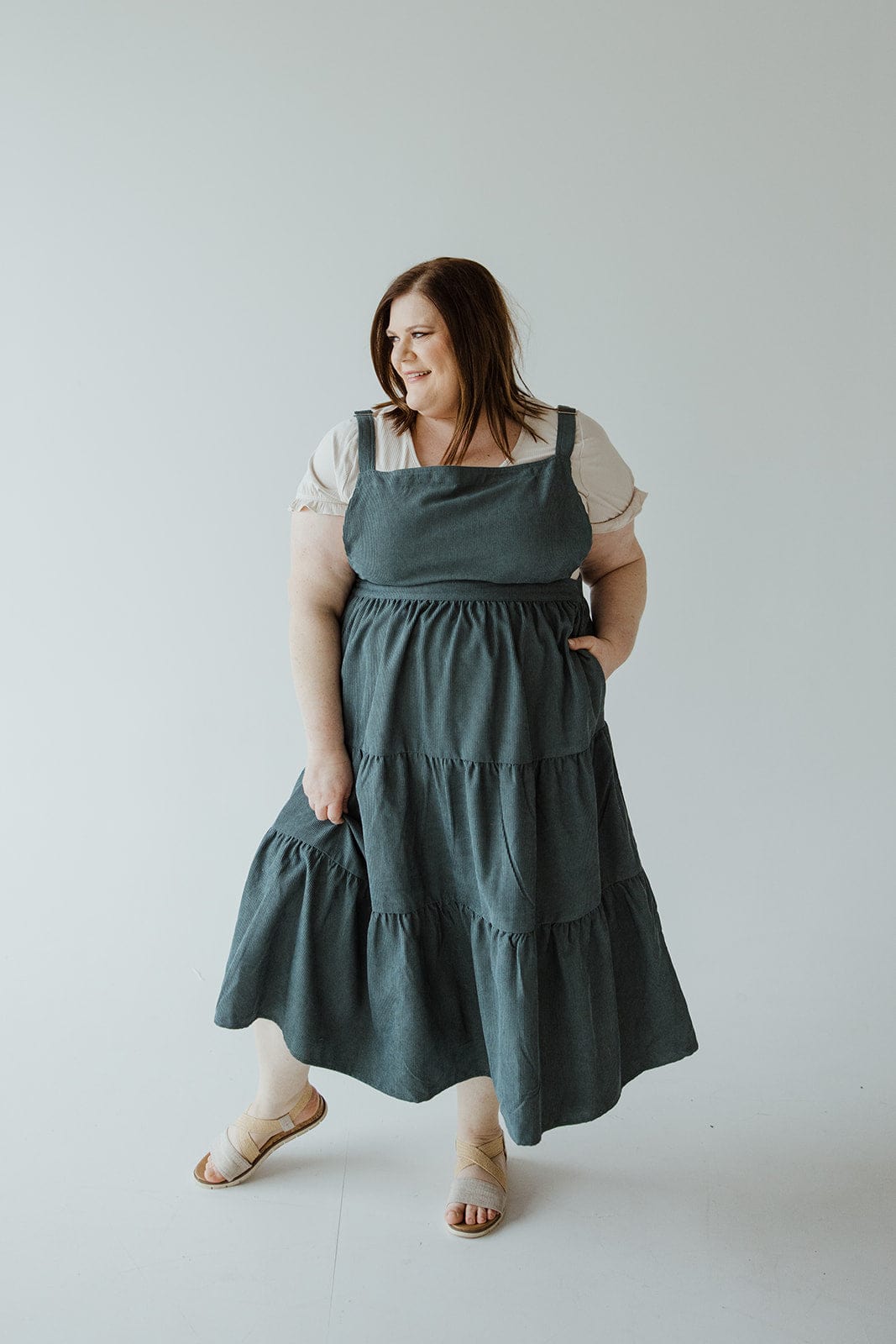 A woman with dark hair wearing a white top and a long, tiered skirt, stands against a plain light grey background. She is looking slightly to her right and has her right hand in her CORDUROY TIERED JUMPER IN BLUE SPRUCE from Love Marlow pocket with an elastic back waist.