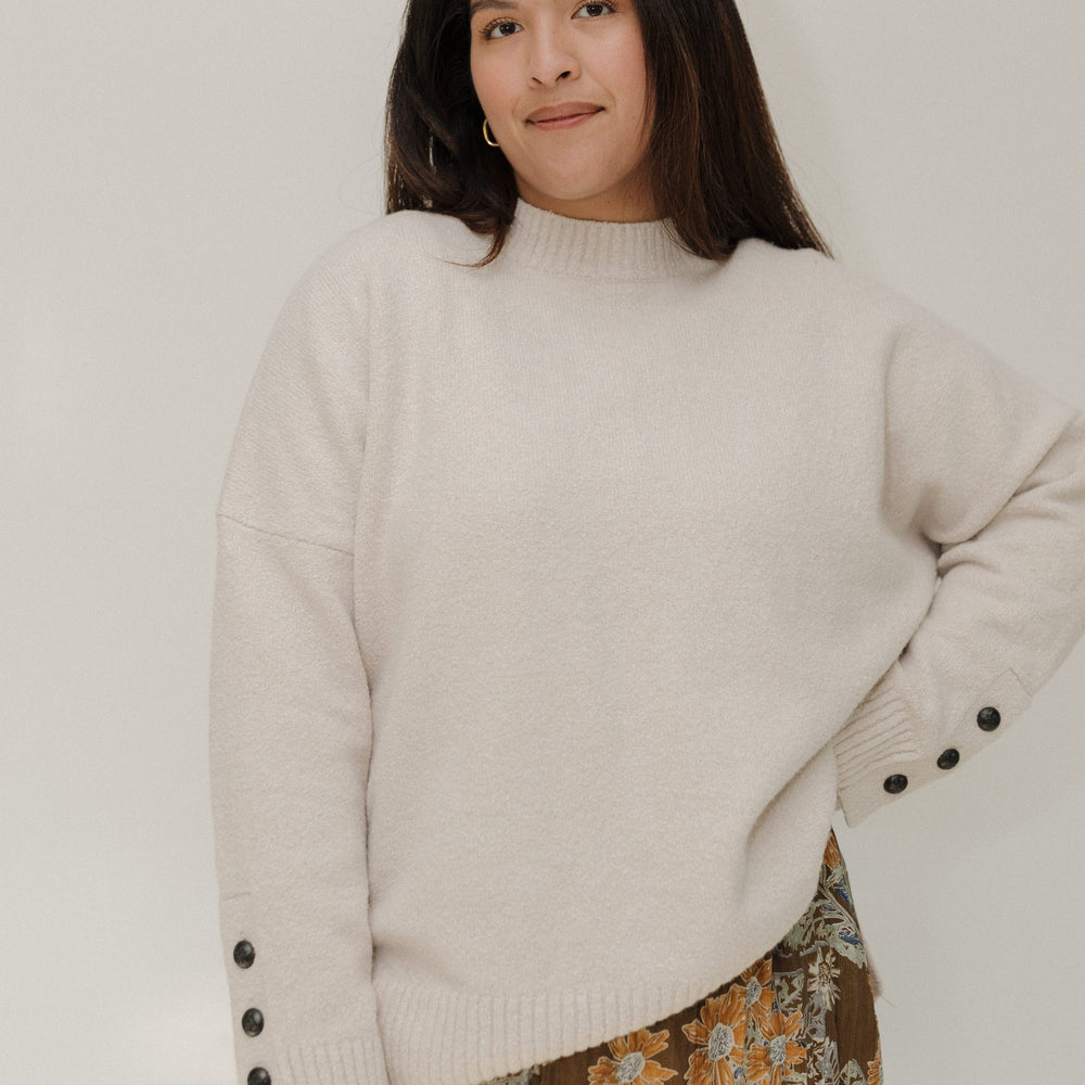 Woman wearing a beige sweater with black buttons and a floral skirt against a white background