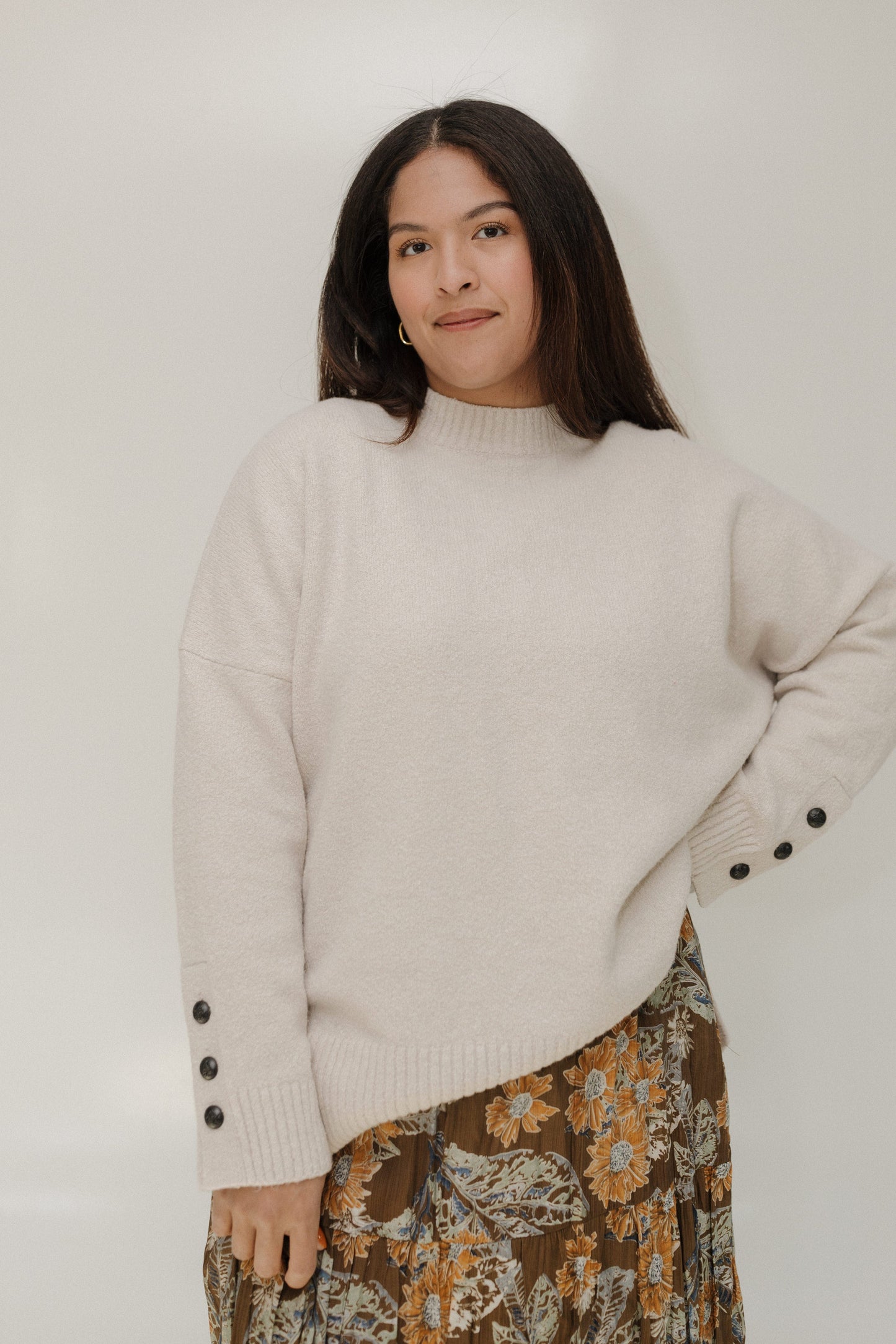 Woman wearing a beige sweater with black buttons and a floral skirt against a white background