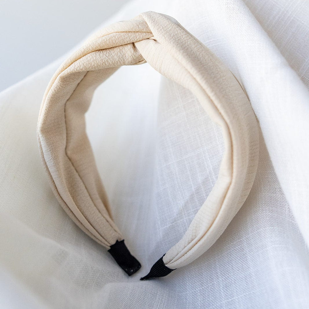 The CRINKLE TEXTURE HEADBAND WITH KNOT IN SAND by Love Marlow is elegantly displayed on a white textured surface.