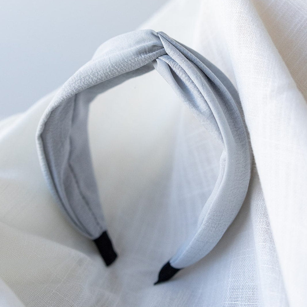 A light gray polyester headband with a twisted design, the CRINKLE TEXTURE HEADBAND WITH KNOT IN STEEL MIST by Love Marlow, rests on a white textured cloth background.