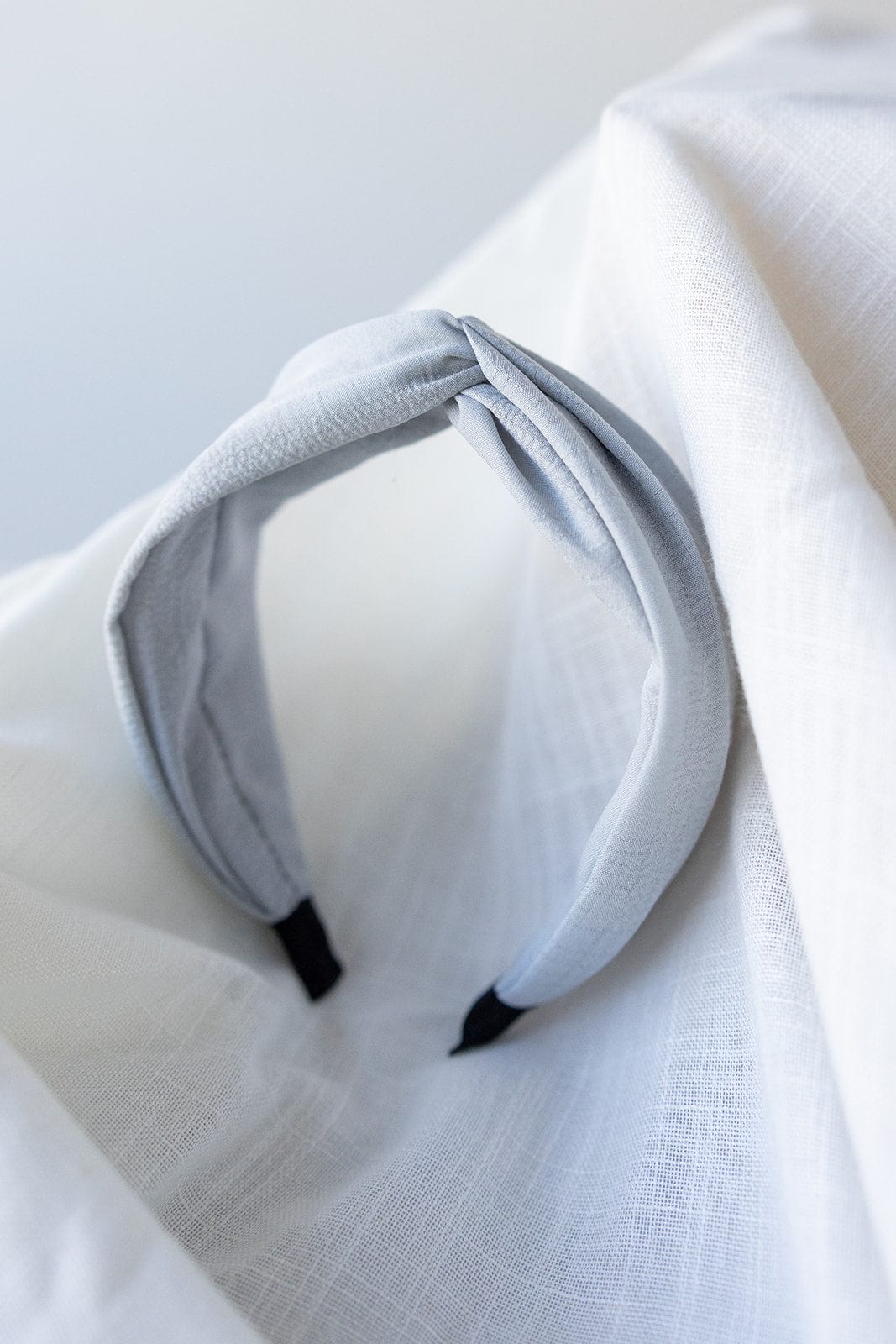 A light gray polyester headband with a twisted design, the CRINKLE TEXTURE HEADBAND WITH KNOT IN STEEL MIST by Love Marlow, rests on a white textured cloth background.