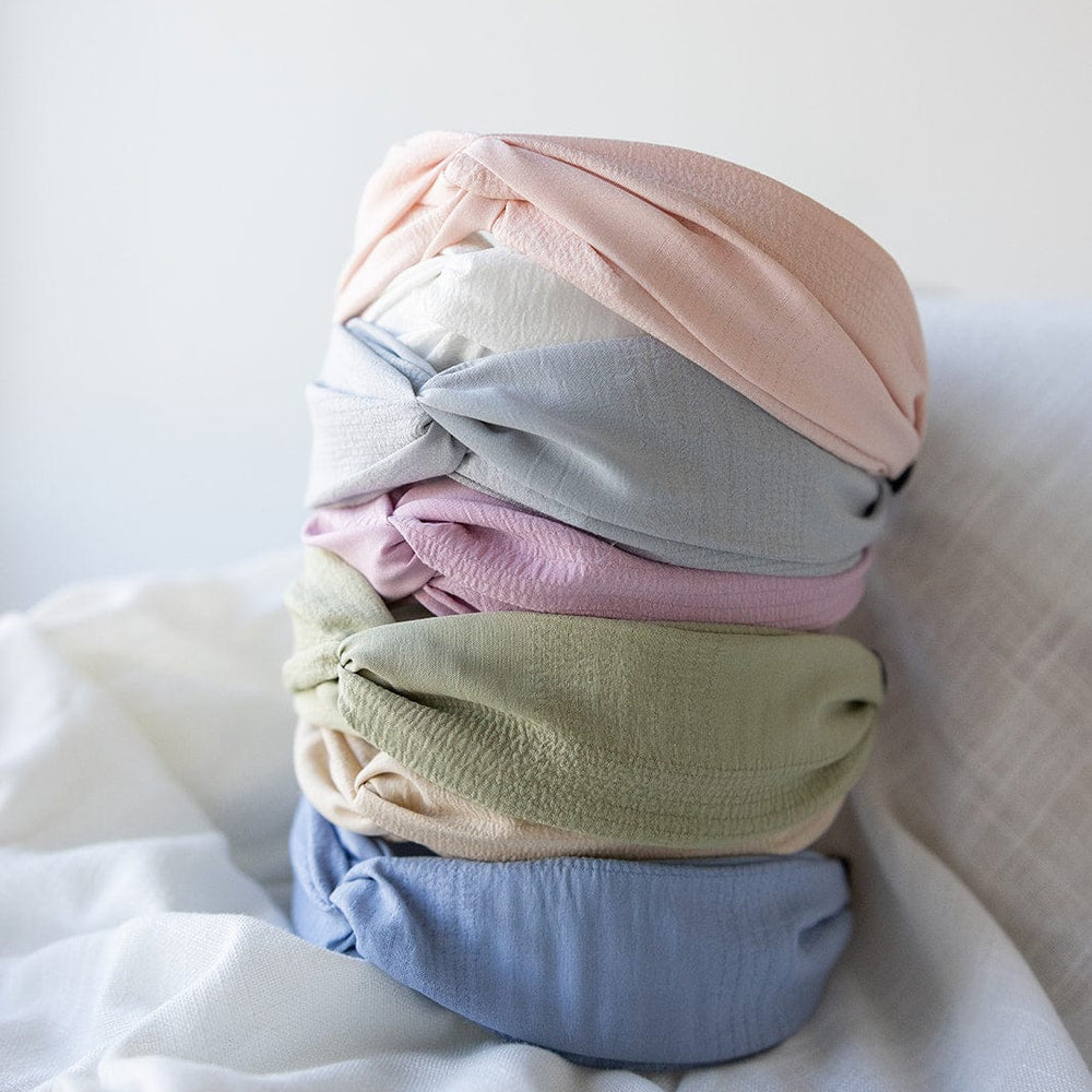 A collection of Love Marlow's CRINKLE TEXTURE HEADBAND WITH KNOT IN STEEL MIST, featuring soft pastel shades including pink, blue, green, beige, and light blue, is neatly stacked on a white fabric surface.