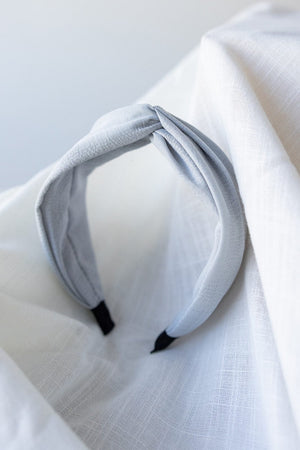 A light gray polyester headband with a twisted design, the CRINKLE TEXTURE HEADBAND WITH KNOT IN STEEL MIST by Love Marlow, rests on a white textured cloth background.