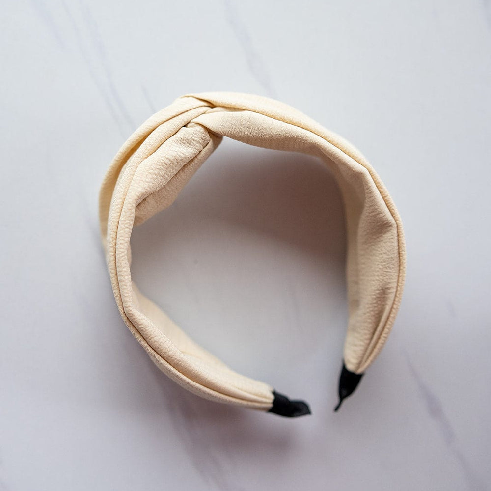 The CRINKLE TEXTURE HEADBAND WITH KNOT IN SAND by Love Marlow, featuring a twisted knot design and black tips, is artfully displayed on a light marbled surface, making it the perfect hair accessory.