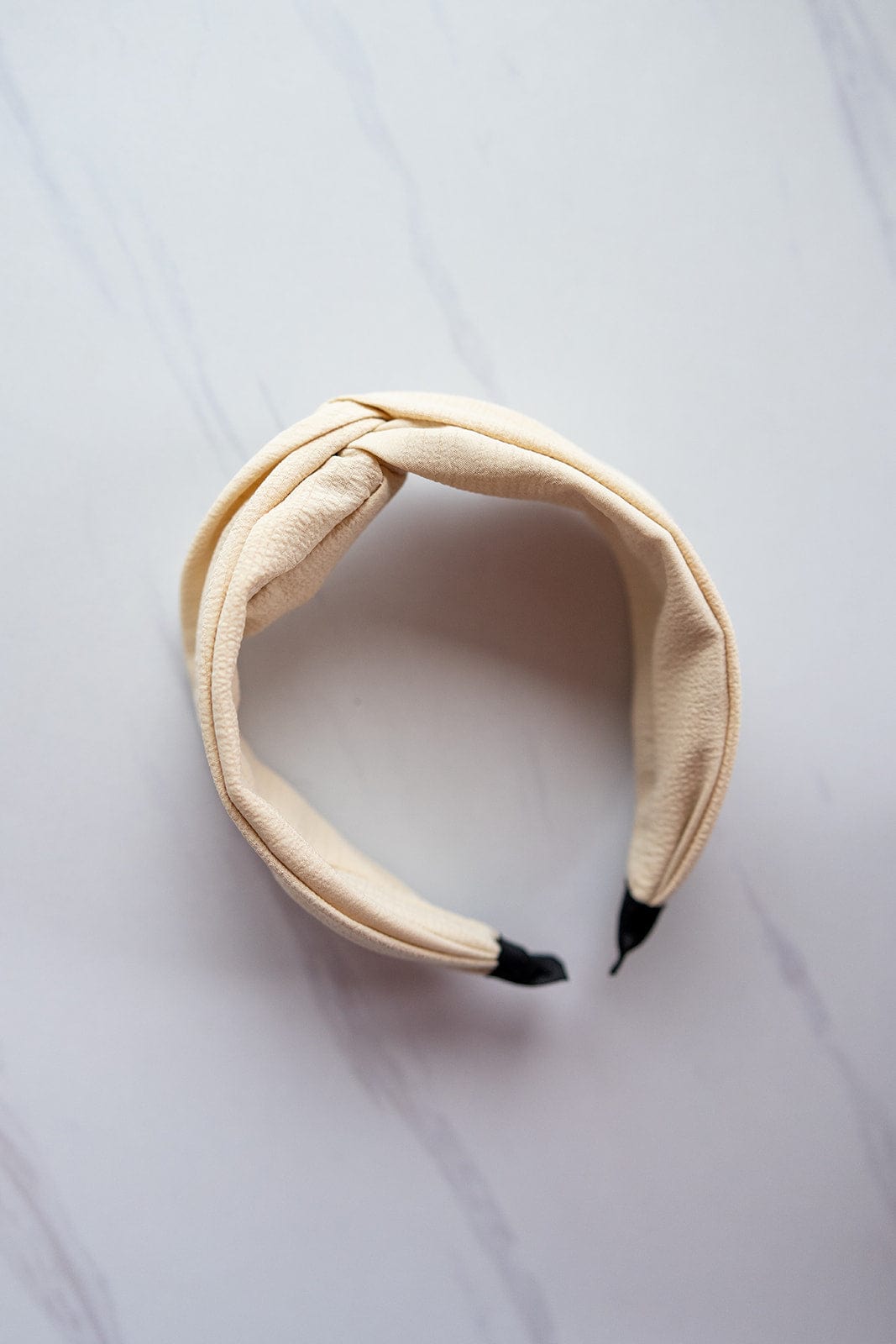 The CRINKLE TEXTURE HEADBAND WITH KNOT IN SAND by Love Marlow, featuring a twisted knot design and black tips, is artfully displayed on a light marbled surface, making it the perfect hair accessory.