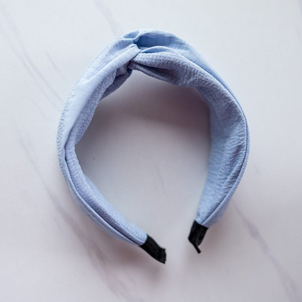 A crinkle texture headband in twilight sky blue features a twisted knot design with black ends, resting elegantly on a white marble background.