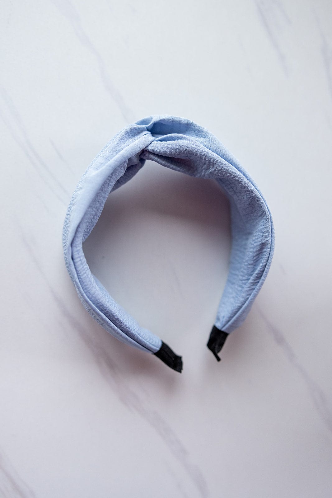 A crinkle texture headband in twilight sky blue features a twisted knot design with black ends, resting elegantly on a white marble background.