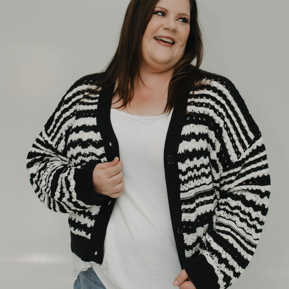 Woman wearing a black and white striped cardigan over a white shirt and blue jeans on a plain background