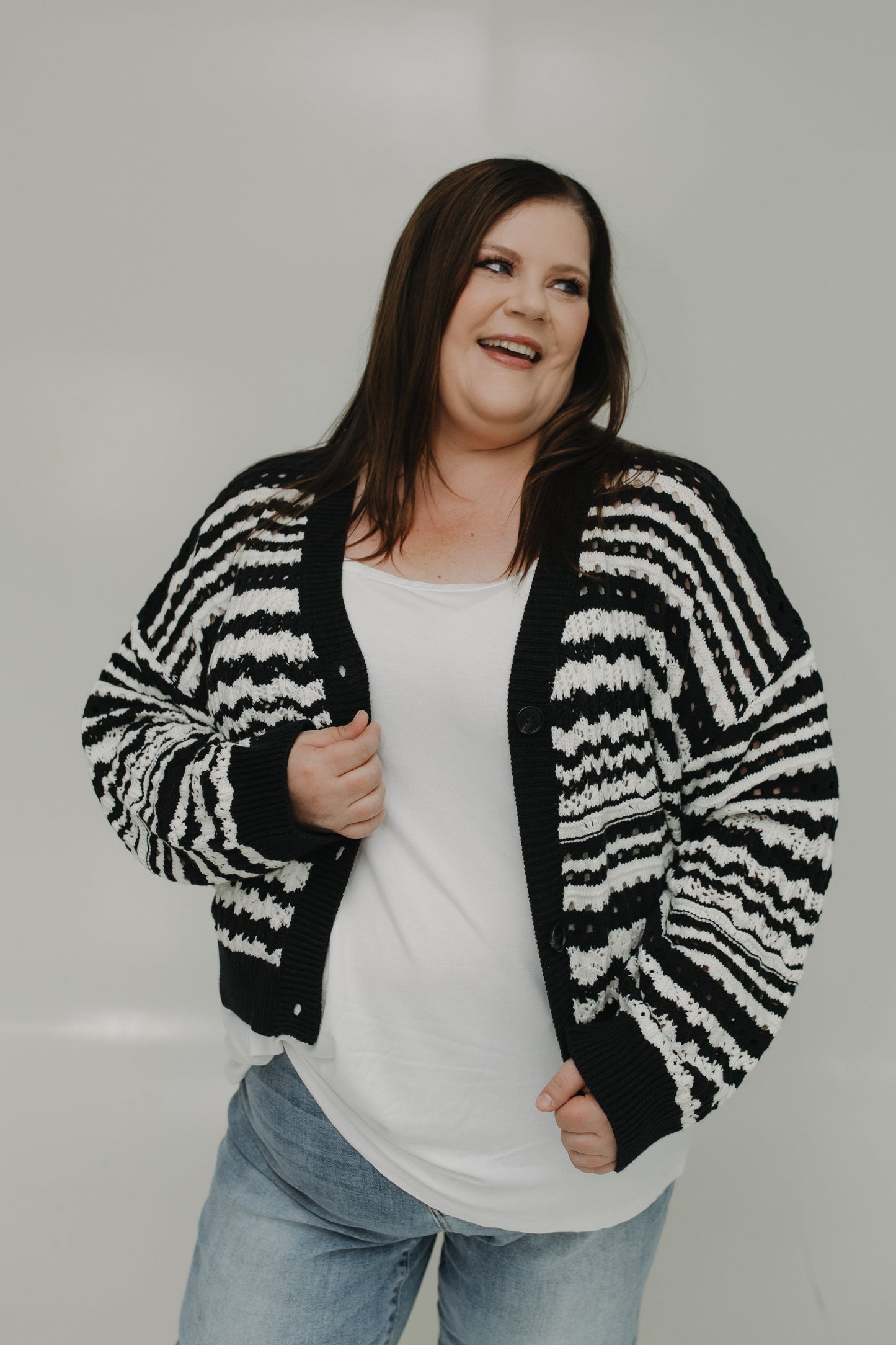 Woman wearing a black and white striped cardigan over a white shirt and blue jeans on a plain background