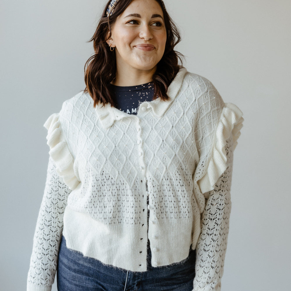 A person wearing a white cardigan with ruffle sleeves, dark jeans, and a headband stands against a plain background.