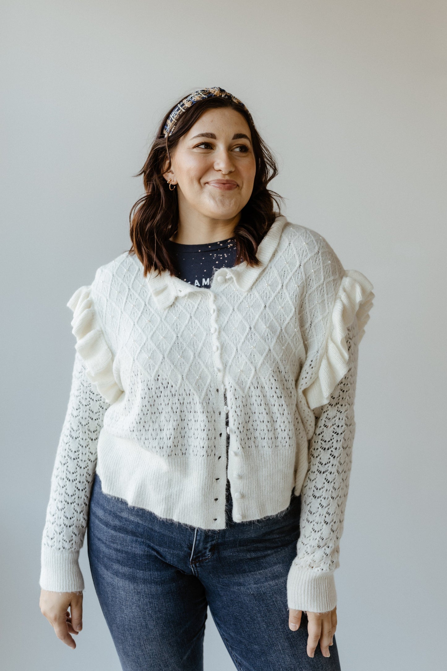 A person wearing a white cardigan with ruffle sleeves, dark jeans, and a headband stands against a plain background.