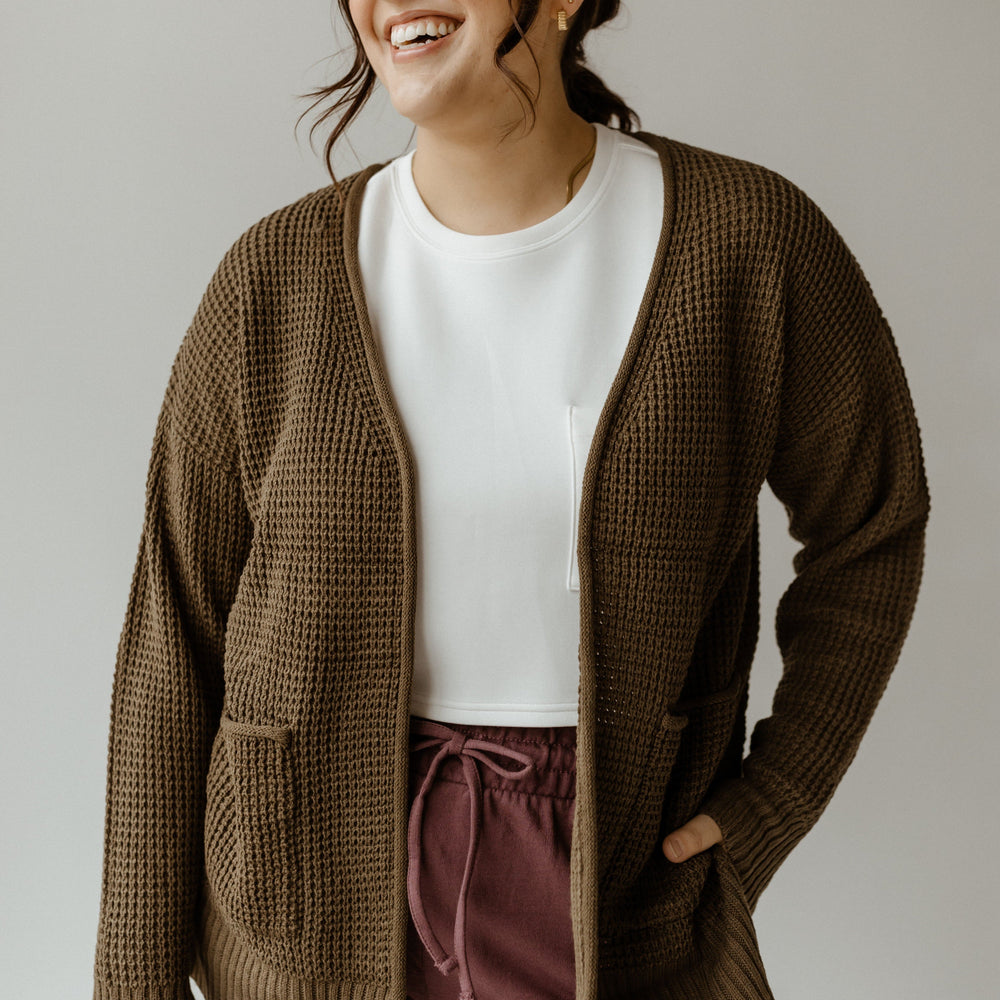 A person smiles while wearing a crochet knit cardigan over a white shirt and purple pants, the antique moss hue beautifully complementing the plain background.