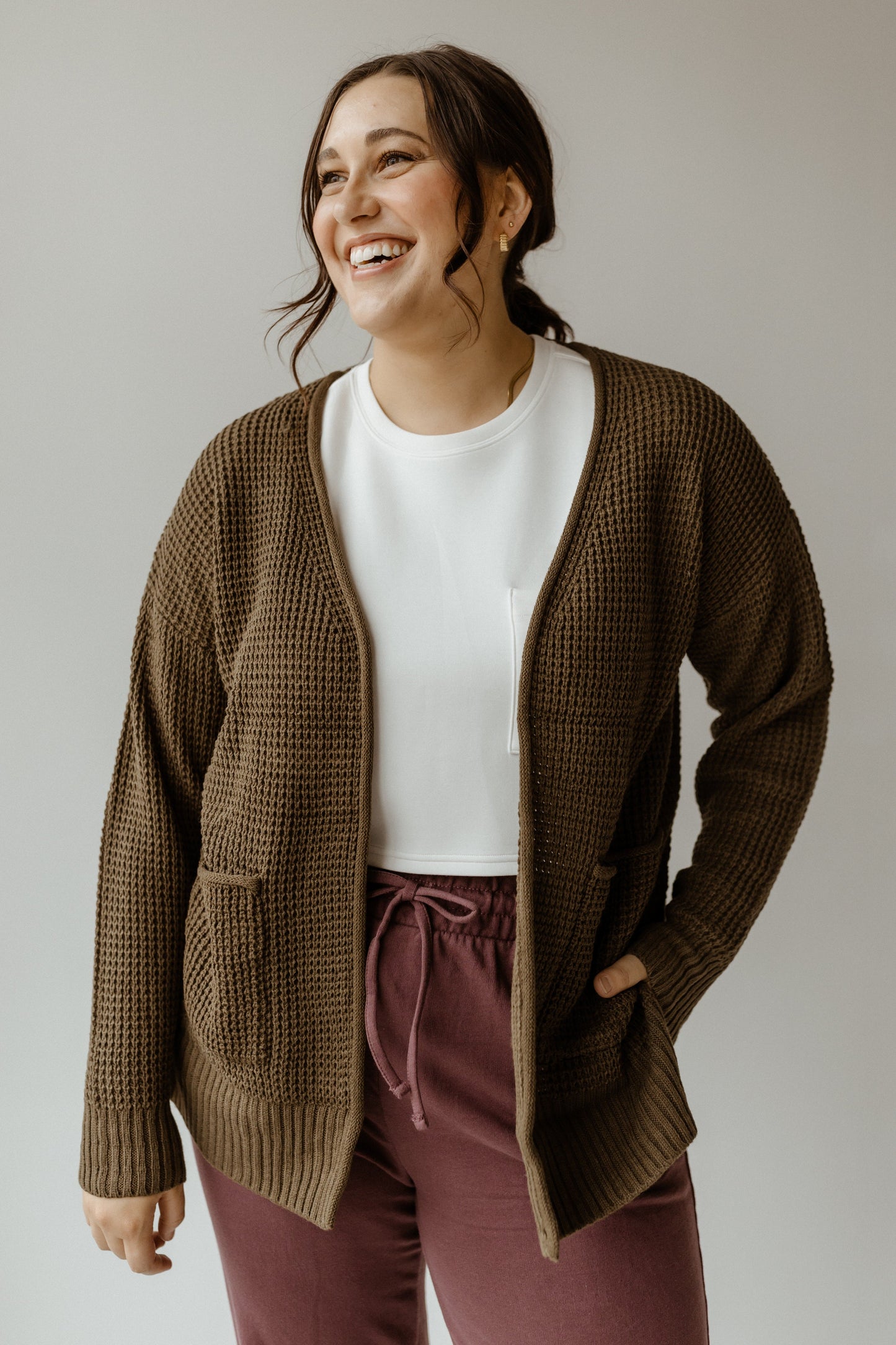 A person smiles while wearing a crochet knit cardigan over a white shirt and purple pants, the antique moss hue beautifully complementing the plain background.