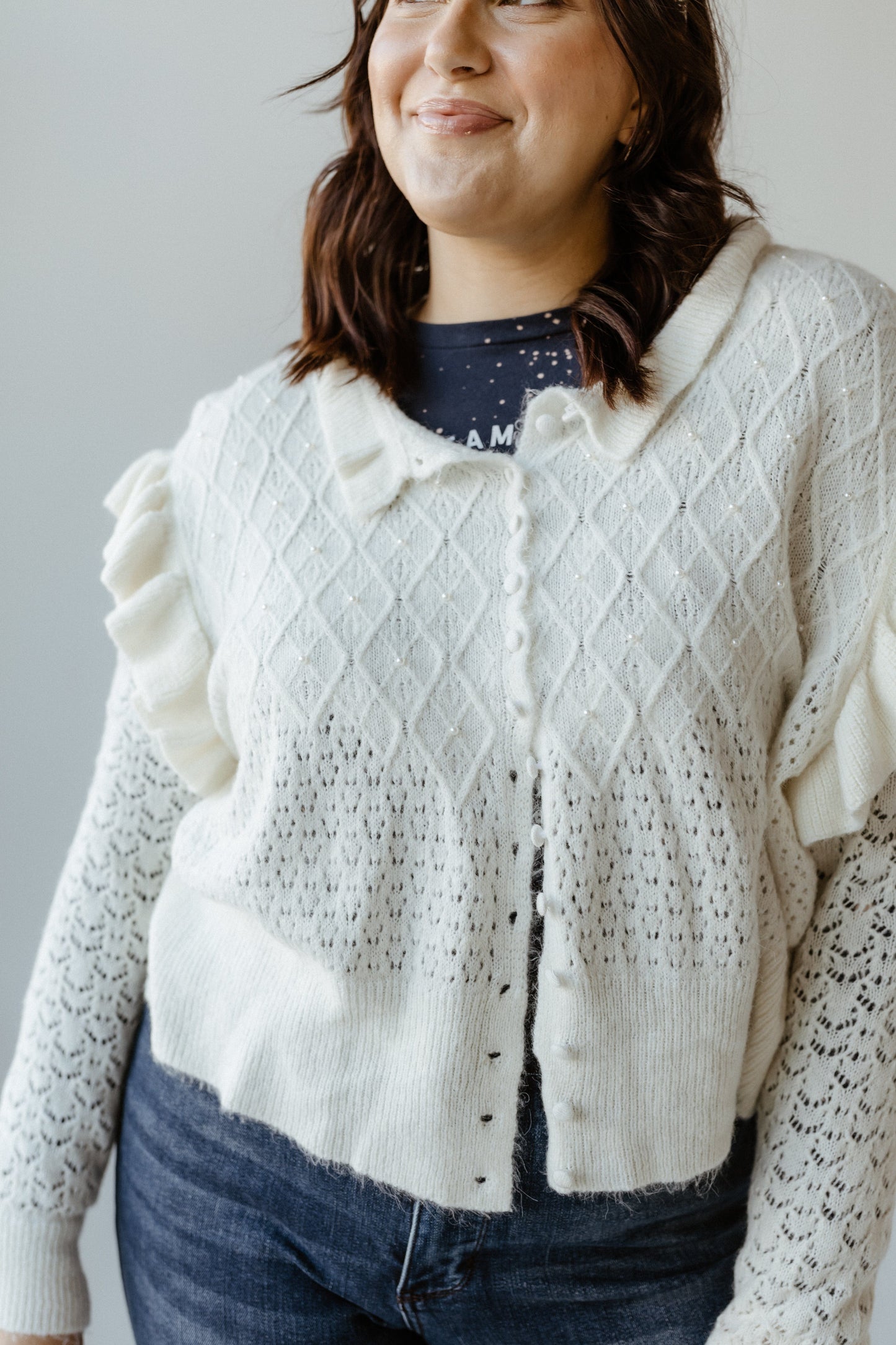 A person wearing a crochet cardigan with pearl embellishments over a dark shirt and blue jeans.