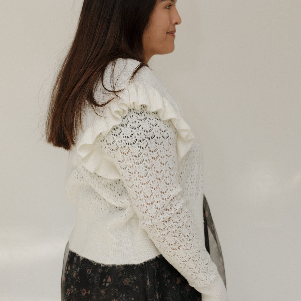 Woman wearing a white lace top and dark floral skirt against a plain background