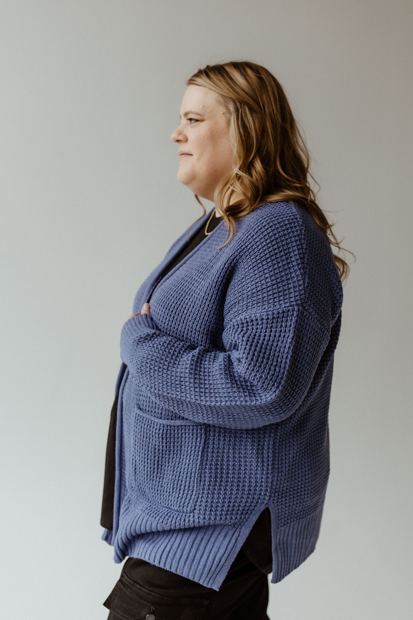 Person with long hair wearing a French Blue crochet knit cardigan and black pants, looking to the side against a plain background.