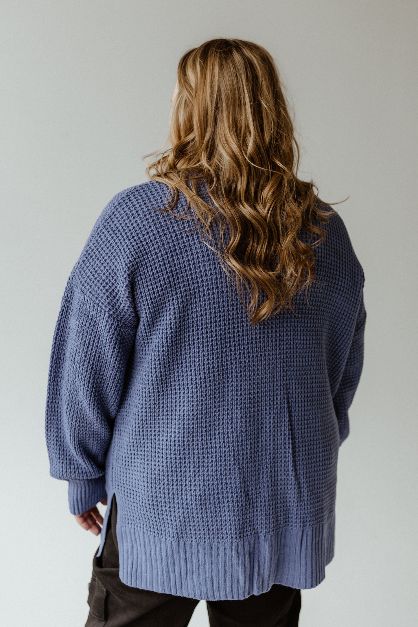 A person with long, wavy hair stands facing away, wearing a French Blue crochet knit cardigan and black pants.