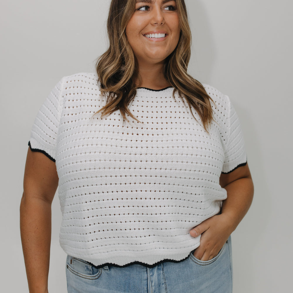 Woman wearing a white crochet top with black trim and blue jeans against a plain background